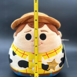 May include: A plush toy of Woody from Toy Story. The toy is approximately 23 cm tall and is wearing his signature cowboy hat, vest, and sheriff's badge. The toy is made of soft, cuddly material and has a friendly expression.