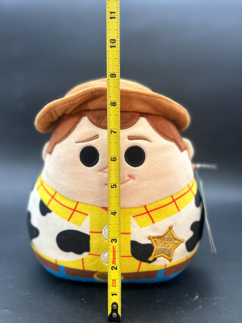 May include: A plush toy of Woody from Toy Story. The toy is approximately 23 cm tall and is wearing his signature cowboy hat, vest, and sheriff's badge. The toy is made of soft, cuddly material and has a friendly expression.