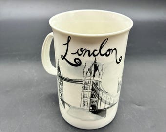 May include: A white ceramic mug with a black and white illustration of the Tower Bridge and the word "London" in cursive. The mug has a handle and a simple design, suitable for beverages.