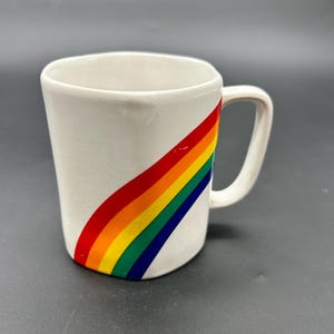 May include: White ceramic mug with a rainbow design. The rainbow is diagonal and has red, orange, yellow, green, blue, and purple stripes.