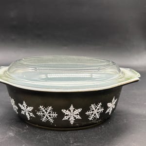 May include: A black Pyrex casserole dish with a clear glass lid. The dish is decorated with white snowflake designs around the rim. A vintage kitchenware item.