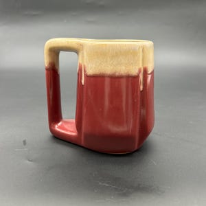 May include: A red and beige ceramic mug with a unique angular design. The mug has a wide, square-shaped handle.