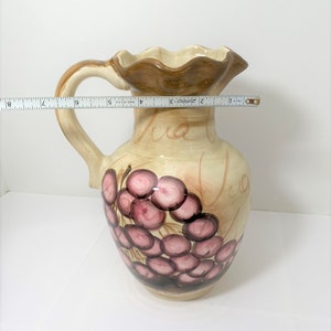 May include: A ceramic pitcher with a brown rim and handle. The pitcher is decorated with a cluster of pink grapes and the words "Via Via" in brown script.