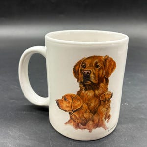May include: A white ceramic mug with a black handle and the text "But First, Coffee" in a bold, black font. The mug is set against a blurred background, suggesting a cozy setting.