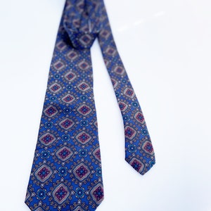 May include: A blue necktie with a repeating geometric pattern in shades of burgundy, gold, and white. The tie is made of a smooth fabric and is displayed against a white background.