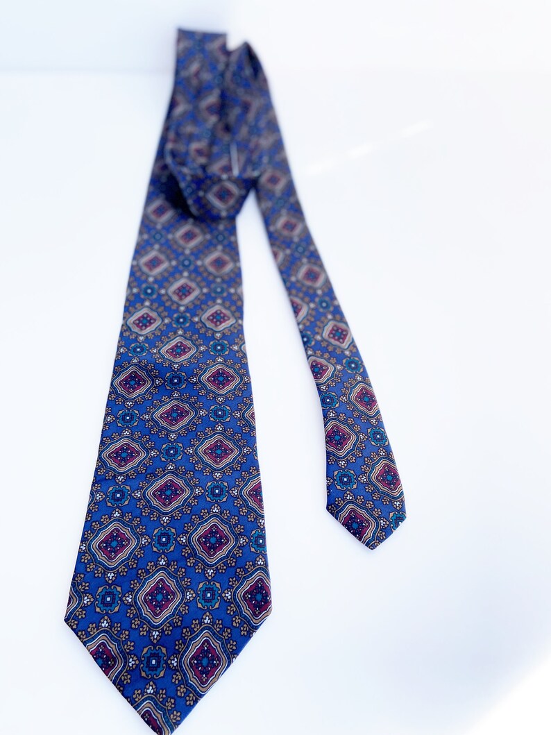 May include: A blue necktie with a repeating geometric pattern in shades of burgundy, gold, and white. The tie is made of a smooth fabric and is displayed against a white background.