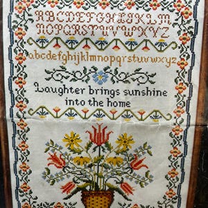Puede incluir: Muestra de punto de cruz con diseños florales y alfabéticos. La muestra presenta el texto « Laughter brings sunshine into the home ». El borde está decorado con flores y vides. La paleta de colores incluye rojo, amarillo, azul y verde.