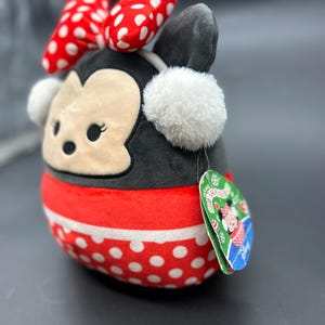 Puede incluir: Un juguete de peluche de un rat&oacute;n de dibujos animados que lleva un vestido de lunares rojos y blancos, un lazo rojo y un gorro de piel blanca con orejeras. El juguete lleva una bufanda a rayas rojas y blancas y una etiqueta que dice "Squishmallows".