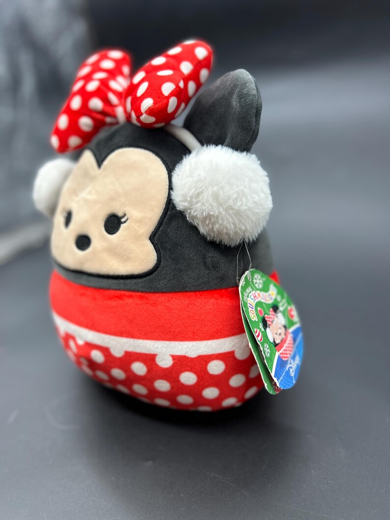 Puede incluir: Un juguete de peluche de un rat&oacute;n de dibujos animados que lleva un vestido de lunares rojos y blancos, un lazo rojo y un gorro de piel blanca con orejeras. El juguete lleva una bufanda a rayas rojas y blancas y una etiqueta que dice "Squishmallows".