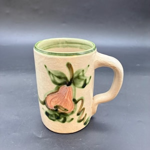 May include: A beige ceramic mug with a hand-painted design of a pear and leaves. The pear is peach-colored, and the leaves are green. The rim of the mug is trimmed with green paint, and it has a curved handle.