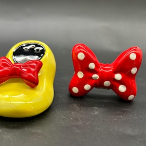 Puede incluir: Un zapato de cerámica amarillo con un lazo rojo y un lazo rojo con lunares blancos. El zapato tiene un interior negro. Probablemente sean objetos decorativos o de colección.