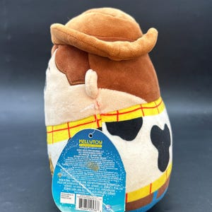 May include: A brown and tan plush toy of Woody from Toy Story. The toy is wearing a brown cowboy hat, a blue shirt, and a yellow vest with a red bandana. The toy has a black and white cow pattern on its chest. The toy is standing on a white background. The toy's tag is visible and reads "KELLYTOY A JAKKS PACIFIC COMPANY".