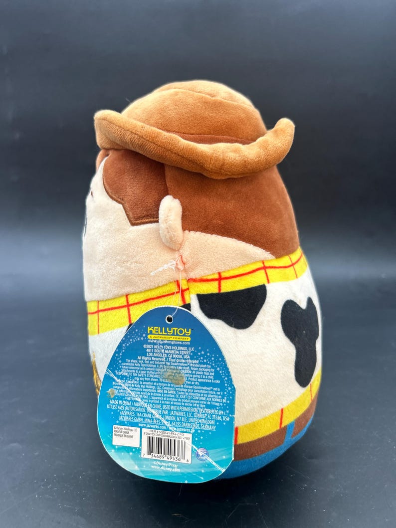 May include: A brown and tan plush toy of Woody from Toy Story. The toy is wearing a brown cowboy hat, a blue shirt, and a yellow vest with a red bandana. The toy has a black and white cow pattern on its chest. The toy is standing on a white background. The toy's tag is visible and reads "KELLYTOY A JAKKS PACIFIC COMPANY".