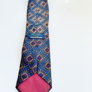 May include: A blue patterned tie with a repeating geometric design in gold, burgundy, and teal. The tie has a burgundy backing and a label that reads "HAND SEWN BY Frazier & Hampson for nordstrom."
