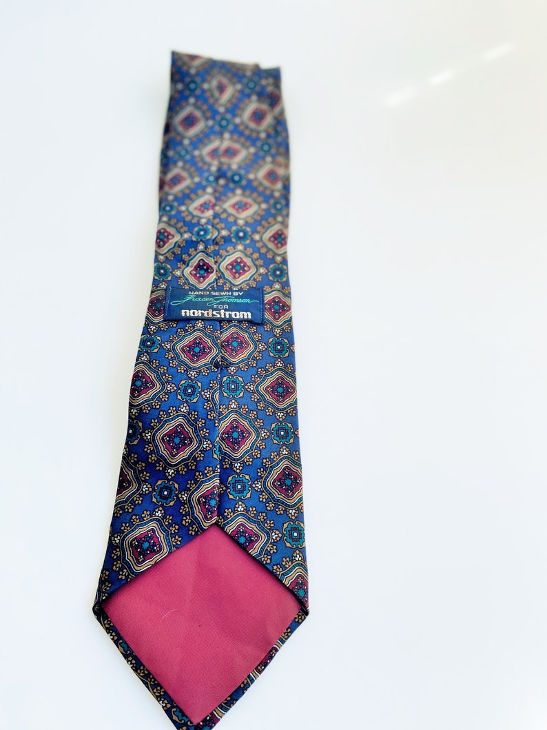May include: A blue patterned tie with a repeating geometric design in gold, burgundy, and teal. The tie has a burgundy backing and a label that reads "HAND SEWN BY Frazier & Hampson for nordstrom."