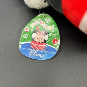 Puede incluir: Una etiqueta verde y azul con las palabras "Original Squishmallows" y "Minnie Mouse" en ella. La etiqueta presenta una imagen de dibujos animados de Minnie Mouse con un vestido de lunares rojos y blancos y un lazo rojo. La etiqueta tambi&eacute;n tiene un fondo azul con un logotipo de Disney blanco.