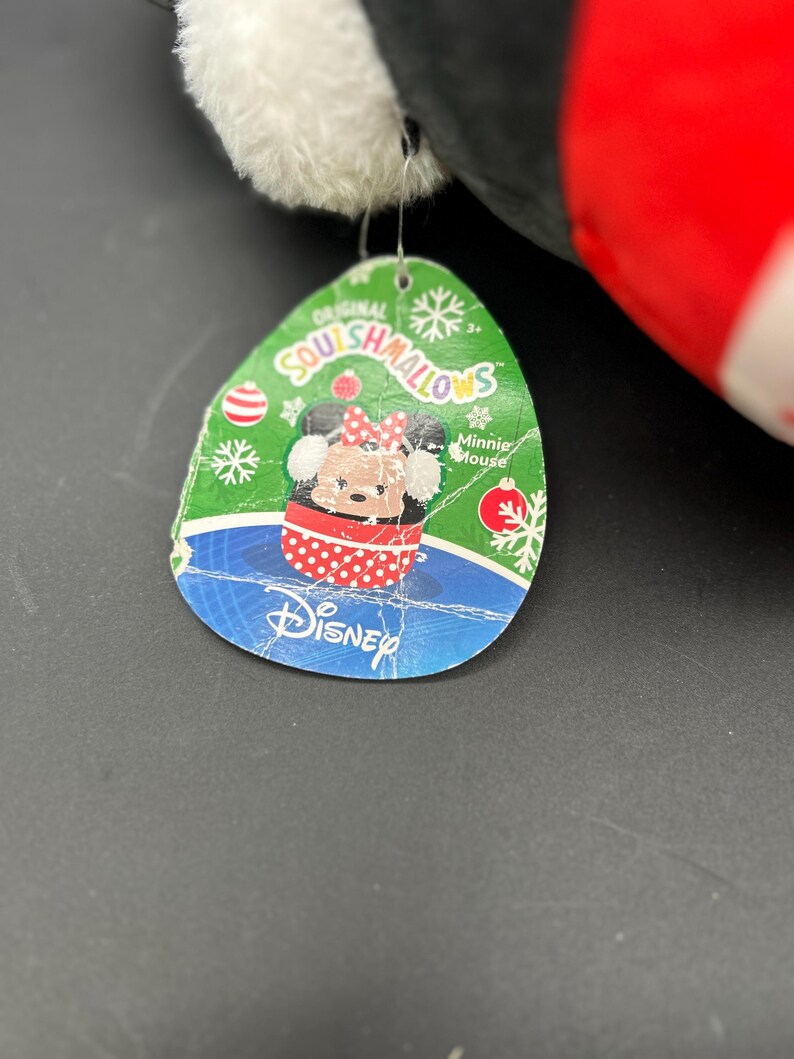 Puede incluir: Una etiqueta verde y azul con las palabras "Original Squishmallows" y "Minnie Mouse" en ella. La etiqueta presenta una imagen de dibujos animados de Minnie Mouse con un vestido de lunares rojos y blancos y un lazo rojo. La etiqueta tambi&eacute;n tiene un fondo azul con un logotipo de Disney blanco.