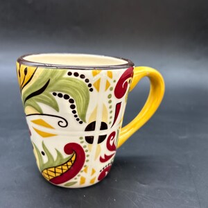 Puede incluir: Una taza de cerámica con asa amarilla y cuerpo color crema. La taza está decorada con un colorido estampado floral en verde, rojo, amarillo y negro. El borde de la taza es marrón.