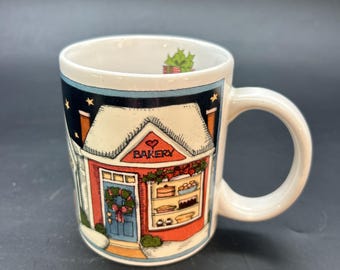 Pode incluir: Caneca de cerâmica branca com uma ilustração de uma padaria. A padaria tem uma fachada vermelha, uma porta azul e um telhado coberto de neve. A palavra "Bakery" está acima da janela. A caneca tem uma alça curva.