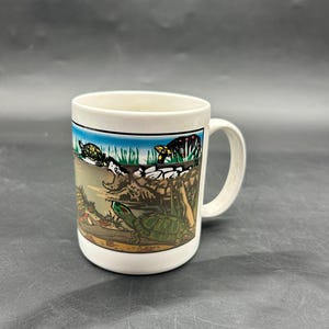 May include: White ceramic mug featuring a colorful illustration of turtles in a pond. The design includes green, brown, and blue hues, with a black border at the top. The mug has a curved handle.