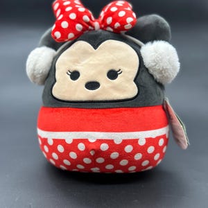 Puede incluir: Un juguete de peluche con forma de rat&oacute;n de dibujos animados que lleva un lazo rojo con lunares blancos y una diadema blanca con pompones. El juguete es gris con un patr&oacute;n de lunares rojos y blancos en la parte inferior.