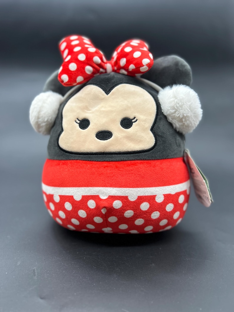 Puede incluir: Un juguete de peluche con forma de rat&oacute;n de dibujos animados que lleva un lazo rojo con lunares blancos y una diadema blanca con pompones. El juguete es gris con un patr&oacute;n de lunares rojos y blancos en la parte inferior.