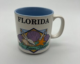 Puede incluir: Taza de cerámica blanca con interior azul. La taza tiene un gráfico de conchas marinas y una estrella de mar con el texto "Florida I've Been There" en una pancarta verde.