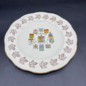 May include: A white decorative plate with a scalloped edge and a gold rim. The plate features brown maple leaf designs around the edge and a central emblem with the word "CANADA" and various coats of arms.