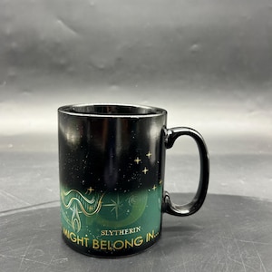 May include: Black ceramic mug with a curved handle. The mug features a green and gold design with the words "Might Belong In... Slytherin" and a star pattern. The mug is cylindrical and has a glossy finish.