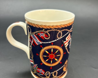 Puede incluir: Una taza de cerámica con temática náutica. La taza presenta un fondo azul marino con imágenes de timones, banderas y detalles de cuerda. El asa y el interior son blancos. La taza es de tamaño estándar para café o té.