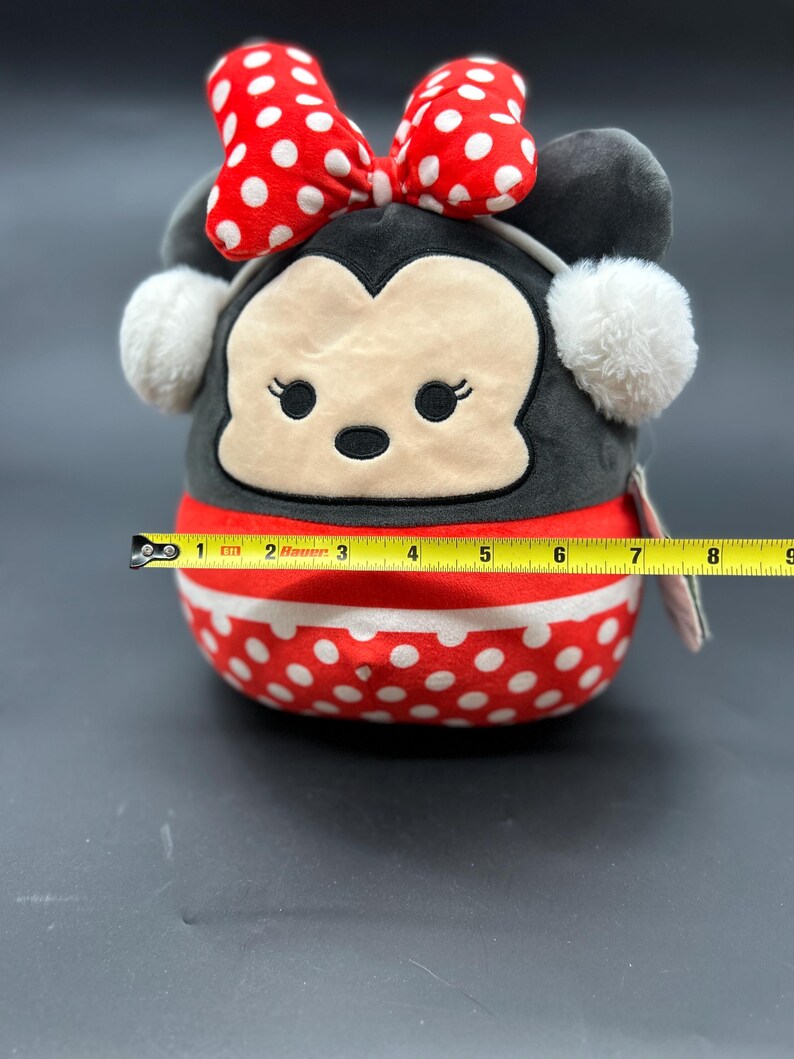 Puede incluir: Un juguete de peluche rojo y blanco con lunares, con una cara negra y un lazo rojo. El juguete tiene orejeras blancas y mide aproximadamente 15 cm de alto.