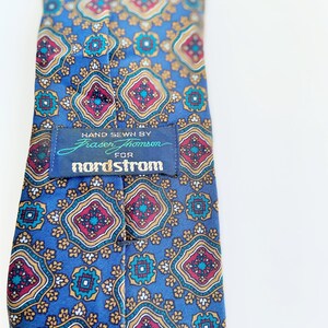 May include: A blue tie with a repeating pattern of red, gold, and teal geometric shapes. The tag reads "HAND SEWN BY Fraser Thomson FOR nordstrom". The tie is a classic accessory.