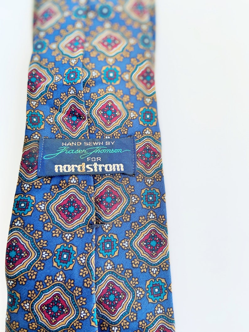 May include: A blue tie with a repeating pattern of red, gold, and teal geometric shapes. The tag reads "HAND SEWN BY Fraser Thomson FOR nordstrom". The tie is a classic accessory.