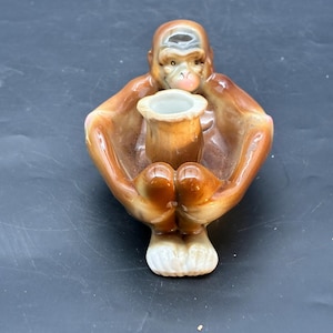 May include: A ceramic monkey figurine in shades of brown and tan, holding a small, cylindrical container. The monkey's arms and legs are outstretched, and its face has detailed features. The base is white.