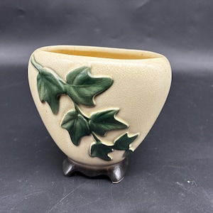 Puede incluir: Jarrón de cerámica color crema con acabado texturizado y base marrón oscuro. El jarrón presenta un diseño de hojas de hiedra verde en relieve en la parte delantera. El jarrón ovalado es un objeto decorativo.