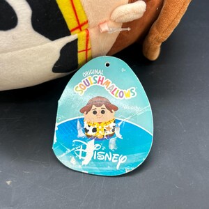 May include: A plush toy of Woody from Toy Story, in the style of a Squishmallow. The toy is white with black spots and a brown cowboy hat. The tag reads "Original Squishmallows" with a cartoon image of Woody and the text "Woody" and "Disney".