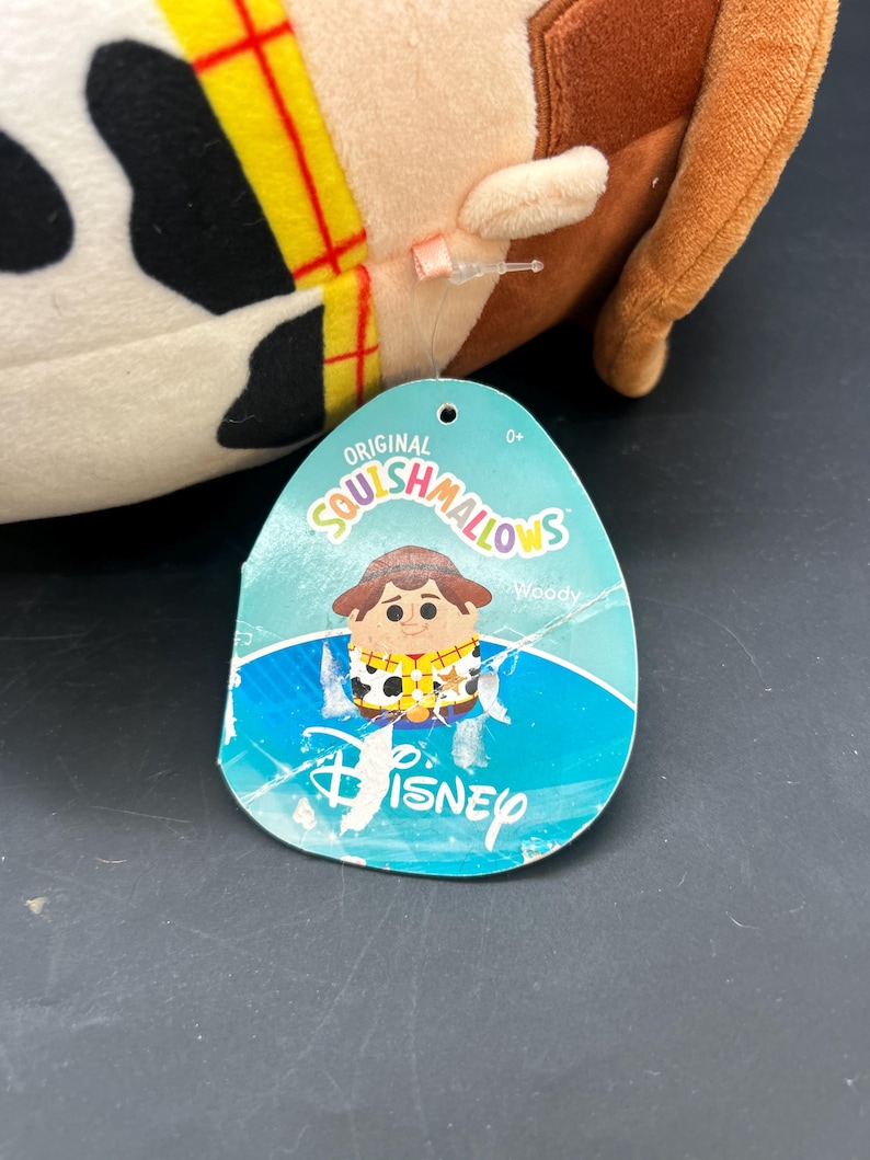 May include: A plush toy of Woody from Toy Story, in the style of a Squishmallow. The toy is white with black spots and a brown cowboy hat. The tag reads "Original Squishmallows" with a cartoon image of Woody and the text "Woody" and "Disney".