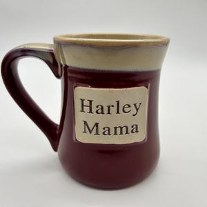 May include: A burgundy ceramic mug with a beige rim and a beige label that reads "Harley Mama".