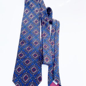 May include: A blue patterned tie with a repeating geometric design in shades of burgundy, teal, and gold. The tie is made of a silky material and is displayed against a white background. The tie is a classic accessory for formal or business attire.