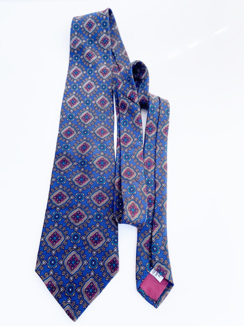 May include: A blue patterned tie with a repeating geometric design in shades of burgundy, teal, and gold. The tie is made of a silky material and is displayed against a white background. The tie is a classic accessory for formal or business attire.
