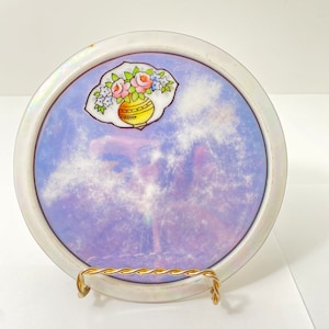 Pode incluir: Um descanso de copo de cerâmica branca com um esmalte iridescente azul e roxo. O descanso de copo apresenta um desenho pintado à mão de um vaso com flores cor-de-rosa, amarelas e verdes.