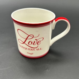 以下が含まれることがあります： 赤い縁と持ち手が付いた白いセラミックマグカップ。「Love conquers all」の文字が赤い筆記体で書かれ、下に「Virgil」と書かれています。シンプルでエレガントなデザインで、日常使いに最適です。