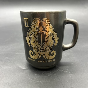 May include: Black ceramic mug with a gold Gemini zodiac design. The mug features the Gemini symbol, two figures, and the text "Gemini May 22 - June 21". The mug has a handle and a slightly tapered shape.