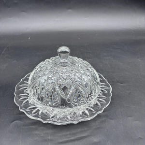 Könnte beinhalten: Eine klare Butterdose aus Glas mit gewölbtem Deckel und dekorativem, gewelltem Rand. Der Deckel hat einen kleinen Knopf zum einfachen Anfassen. Die Schale hat ein rautenförmiges Muster.