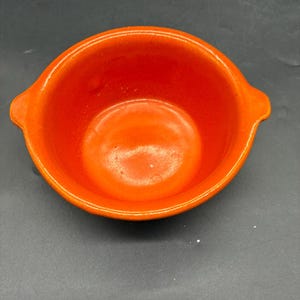 Puede incluir: Cuenco de cerámica naranja brillante con dos pequeñas asas. El cuenco tiene un acabado liso y brillante y una forma redondeada. El interior es del mismo color naranja brillante que el exterior. Este cuenco es probablemente para servir o preparar alimentos. Aproximadamente 15 cm de diámetro.
