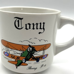 Può includere: Tazza in ceramica bianca con manico. La tazza presenta un'immagine di un biplano stilizzato con il testo "Tony", "U.S. Army" e "Boeing P-12" stampato su di essa.