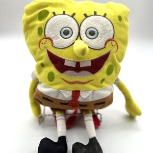 May include: A yellow plush toy of SpongeBob SquarePants sitting with a big smile. He is wearing a brown tie and a white shirt with a brown belt. His eyes are blue and his nose is yellow.