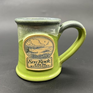 May include: A green and blue ceramic mug with a brown label that reads "Sea Rock Inn Mendocino".