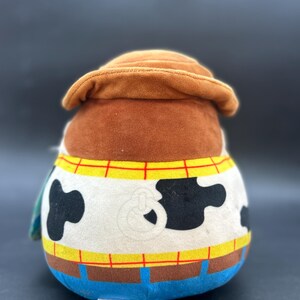 May include: A plush toy of Woody from Toy Story. The toy is brown, white, and blue. Woody is wearing his signature cowboy hat and vest. He has a black and white cow pattern on his shirt.