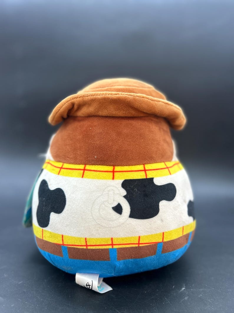 May include: A plush toy of Woody from Toy Story. The toy is brown, white, and blue. Woody is wearing his signature cowboy hat and vest. He has a black and white cow pattern on his shirt.